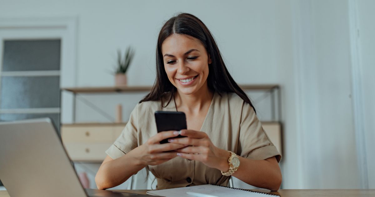 A smiling woman is seated at a modern workspace, setting up her phone hacks on her smartphone in her hands. In the background, a sleek laptop is open, with a notepad and pen set neatly to the side on the desk. The room has a clean, minimalist design with a light color palette, and there's a potted plant that adds a touch of greenery.